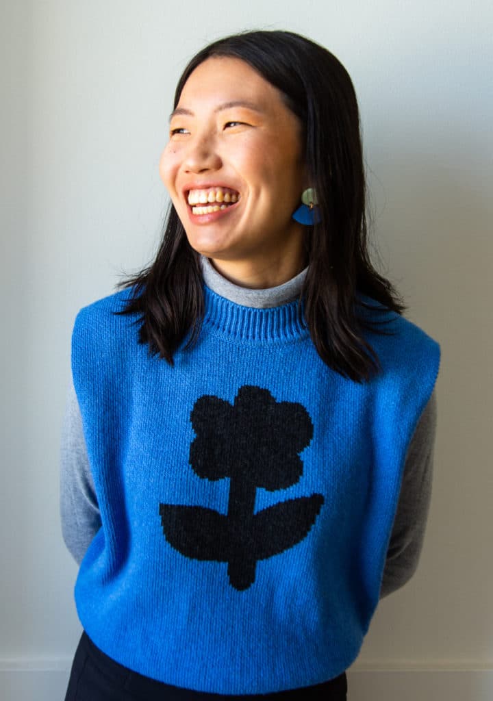 Young woman with black hair in blue sweater vest smiling and looking off in the distance.