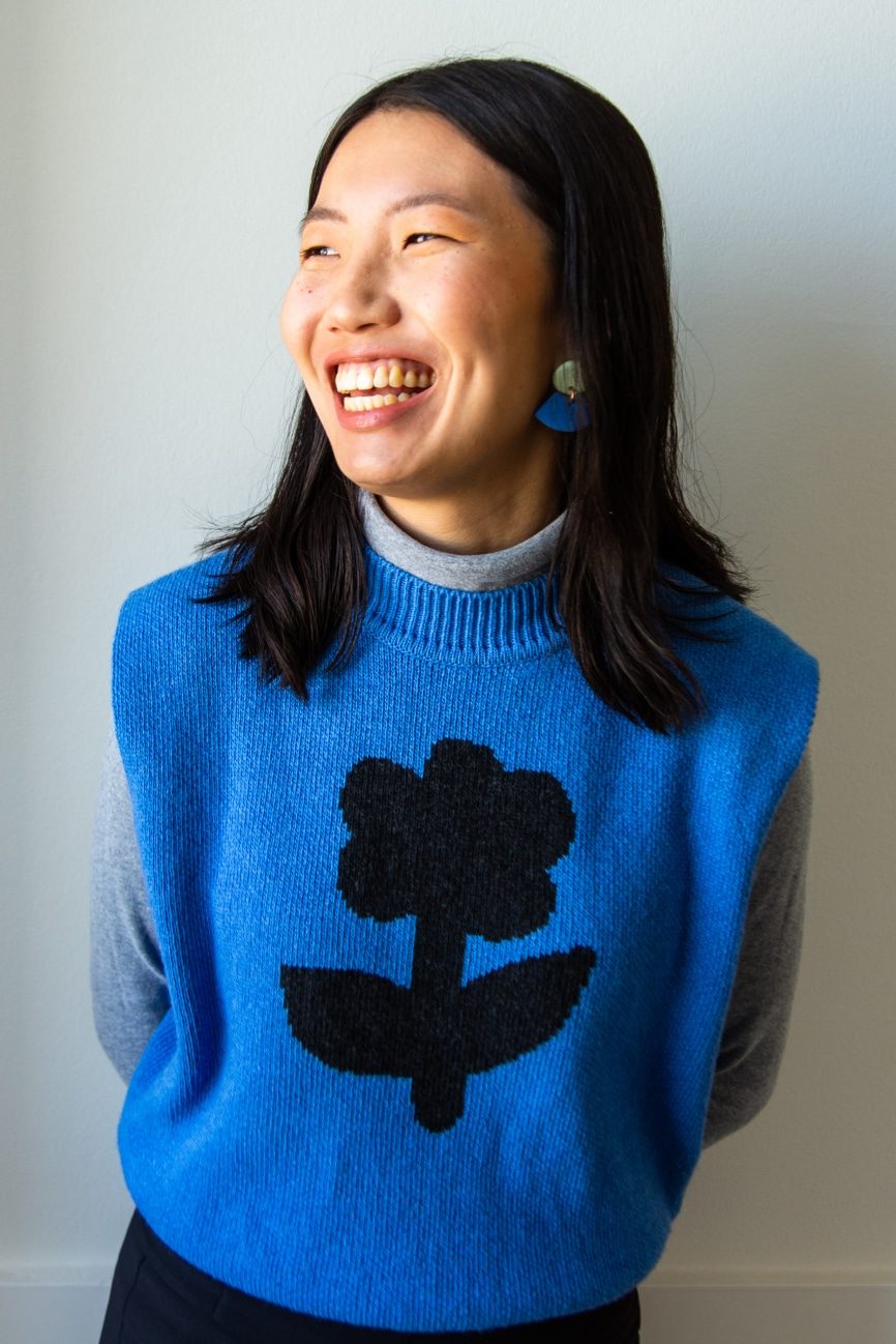 Young woman with black hair in blue sweater vest smiling and looking off in the distance.