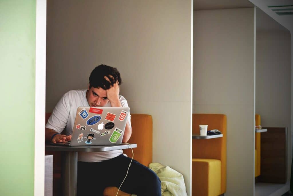 A student sits at a desk looking at a computer screen with his head in his hands. Stickers are on the back of the screen and a coffee cup is on an adjacent table. The pending resumption of student loan repayments will likely stress both individuals and retailers.